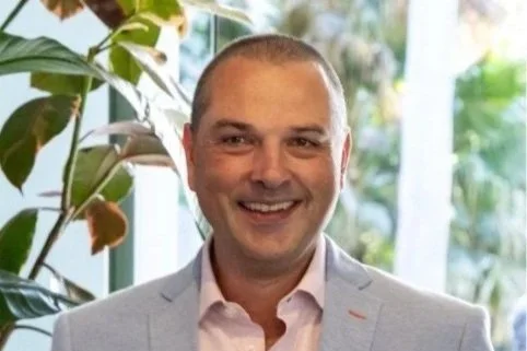 A smiling man in a light gray suit stands in front of lush greenery with a hint of sunlight filtering through, suggesting a vibrant, outdoor setting.