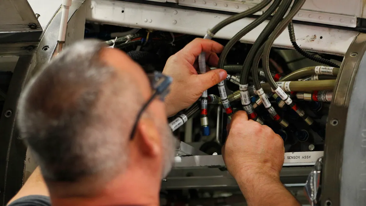 A person with glasses is working inside a machine or electronic system, handling multiple cables and connectors labeled with technical tags.