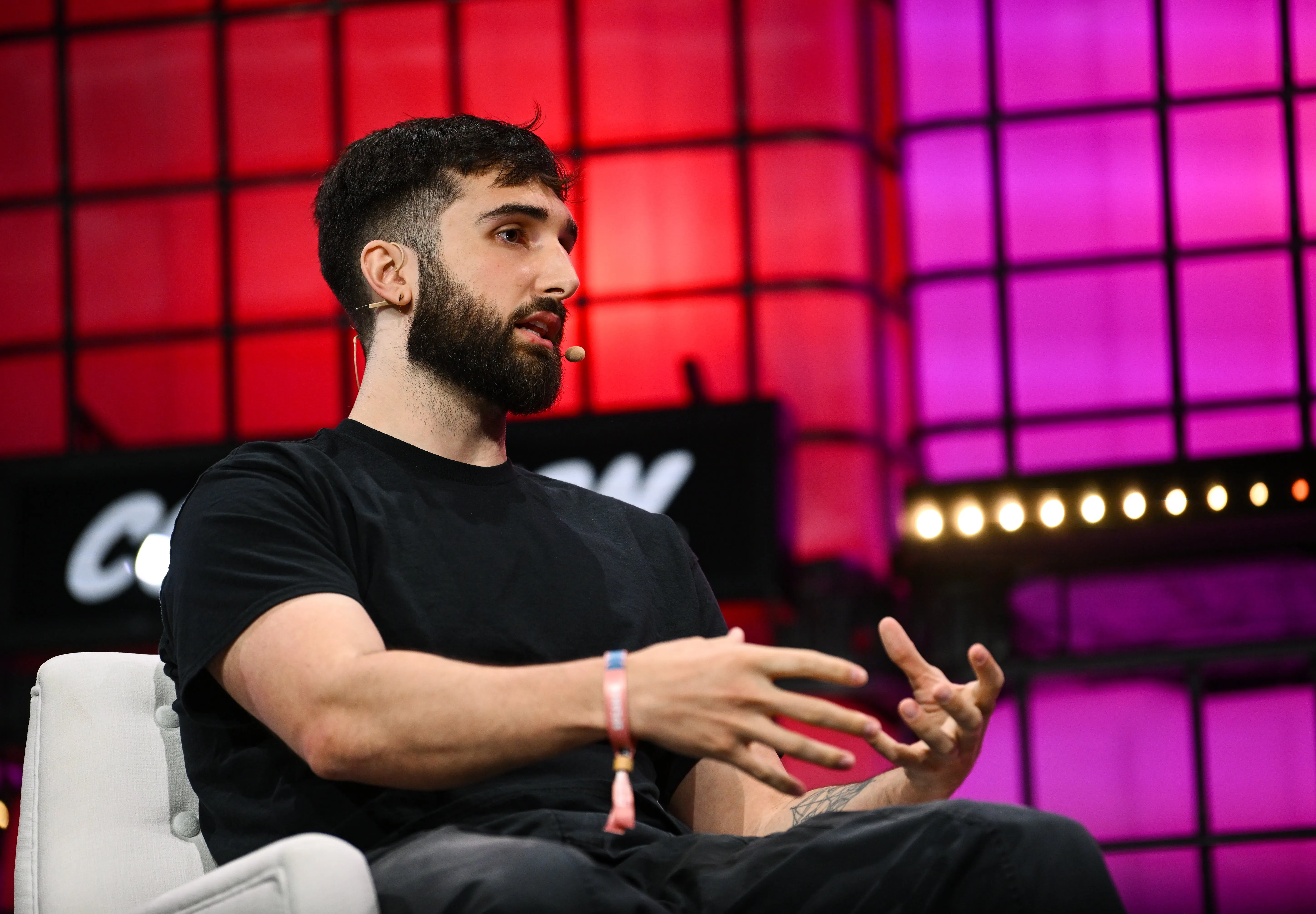 A young man with a beard and dark hair, wearing a black shirt, is speaking or explaining something during a conference or panel discussion, with colorful illumi