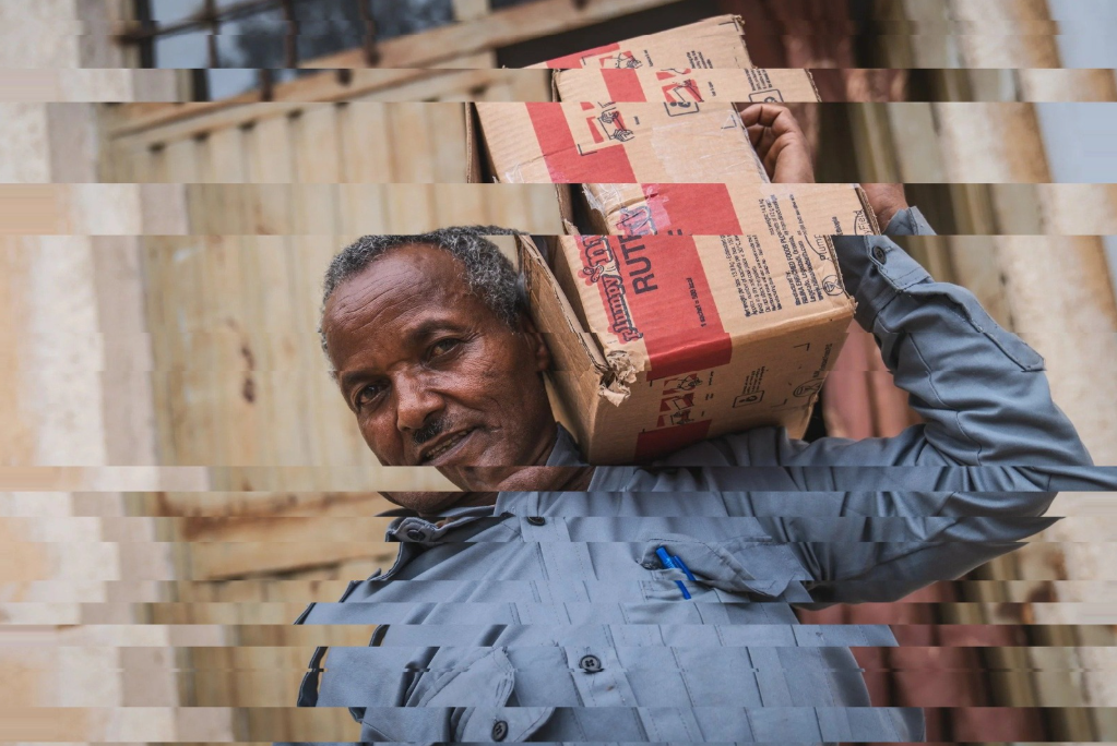 A man in a gray uniform carries a cardboard box on his shoulder, standing in front of a wooden fence.