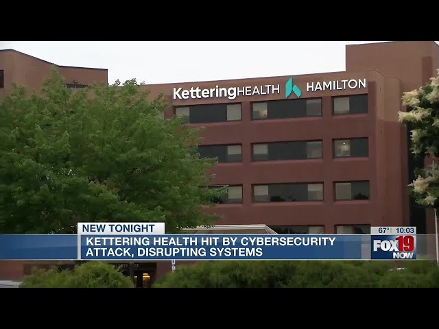 The image shows the Kettering Health building in Hamilton with a news banner reporting a cybersecurity attack disrupting their systems.