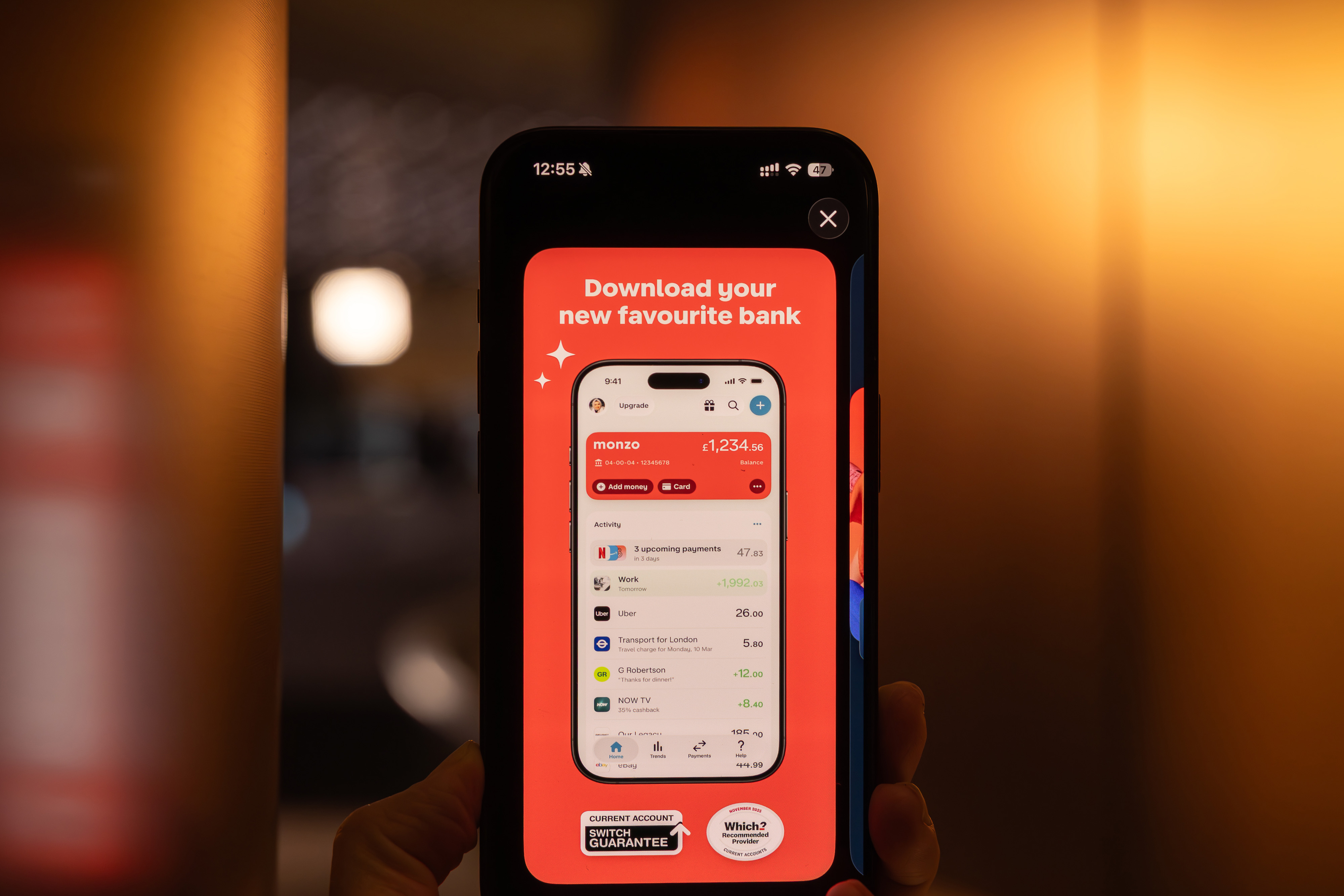 A person holds a smartphone displaying an advertisement for the Monzo bank app, with the text “Download your new favourite bank” and a screenshot of the app’s i