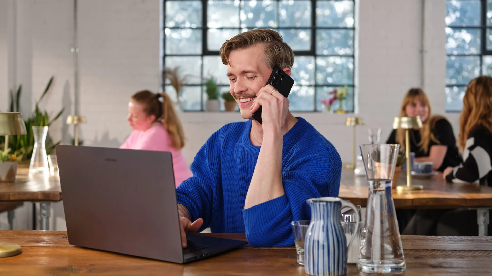 A man smiling while talking on the phone and working on a laptop in a bright, modern office or co-working space with other women in the background.