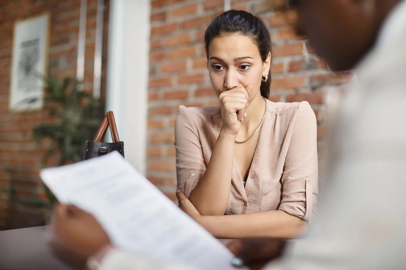 A woman appears worried or anxious while sitting at a table during a job interview, with a person holding a paper in the foreground.